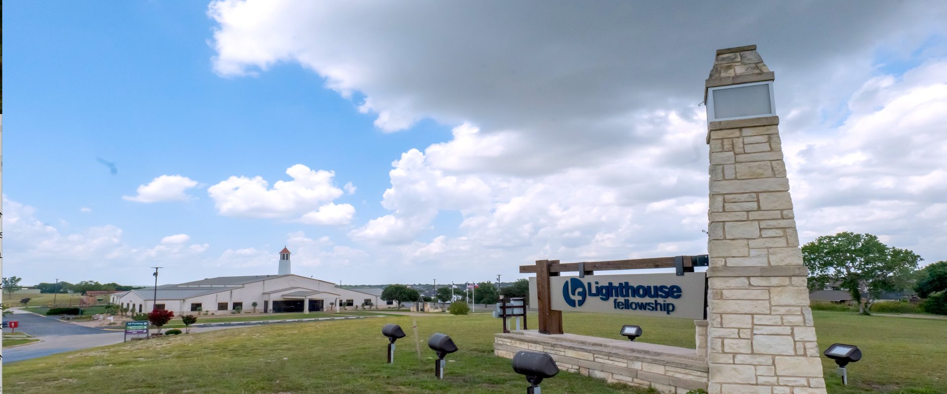 Lighthouse Fellowship church campus exterior with distinctive stone bell tower monument, modern church building, and landscaped grounds under dramatic cloudy sky